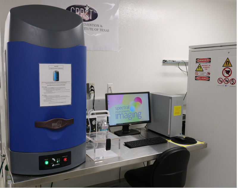 Laboratory setup featuring an Ami HTX Optical Imaging System on the left, a computer monitor displaying 'spectral instruments imaging' in the center, and a white cabinet with multiple safety warning signs on the right.